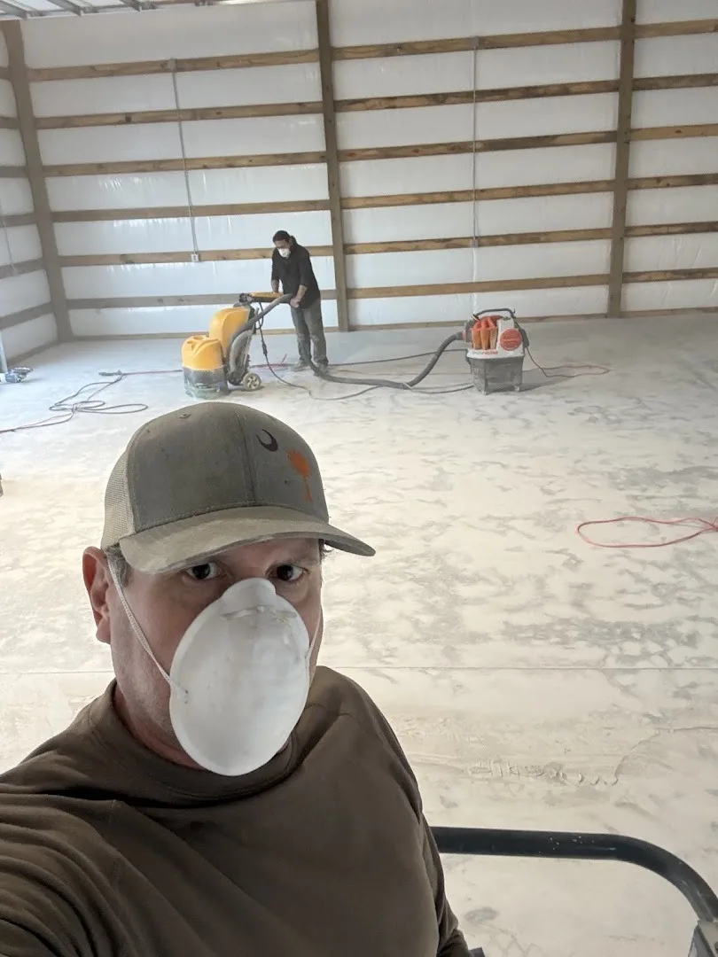 Phil Smith in a dust mask taking a selfie while a floor grinder with a vacuum works on the concrete slab behind him